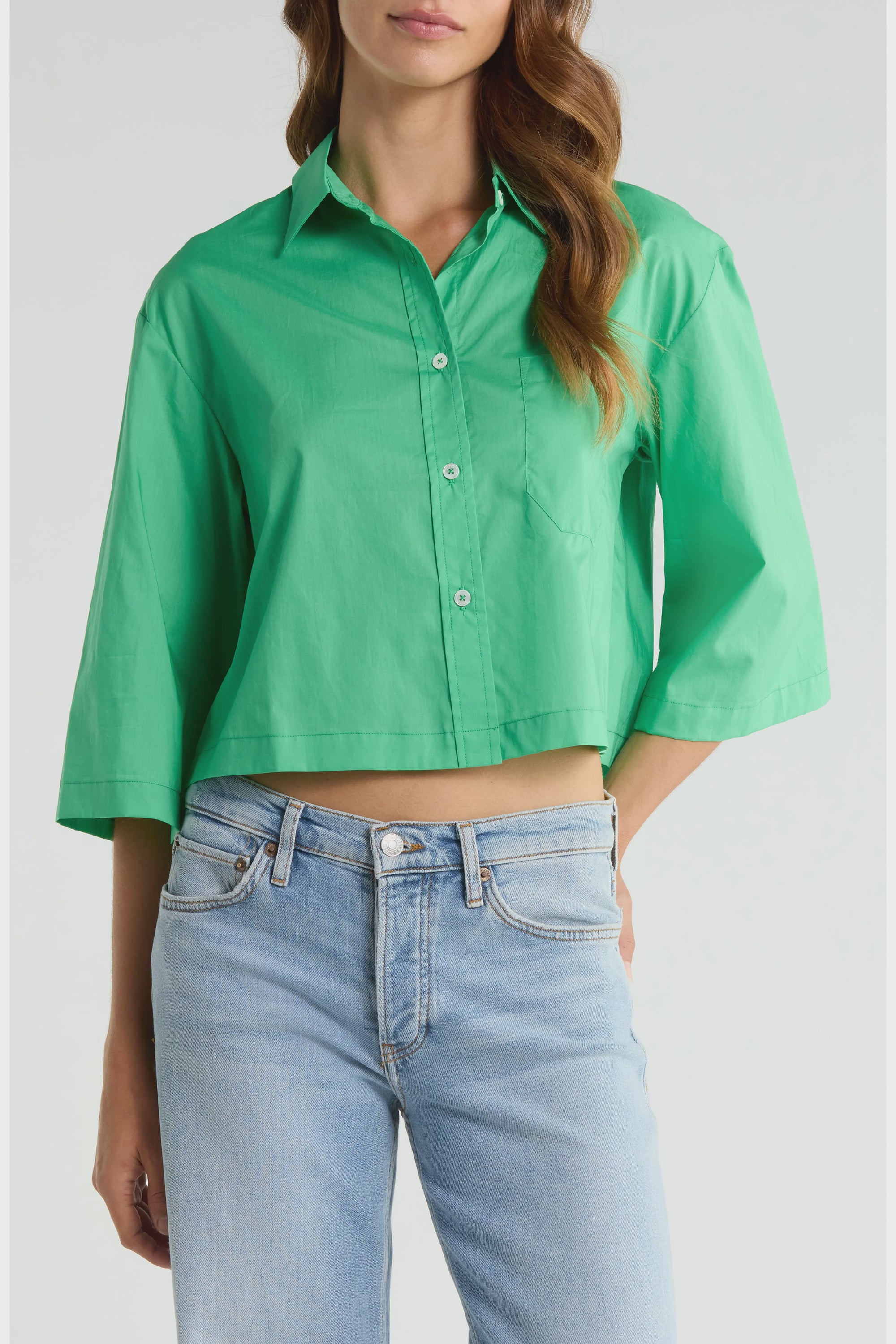 A woman wears The PIXIE Shirt by Cissa, a bright green, boxy button-up with three-quarter sleeves, paired with light blue high-waisted jeans and standing with one hand in her pocket.