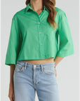 A woman wears The PIXIE Shirt by Cissa, a bright green, boxy button-up with three-quarter sleeves, paired with light blue high-waisted jeans and standing with one hand in her pocket.
