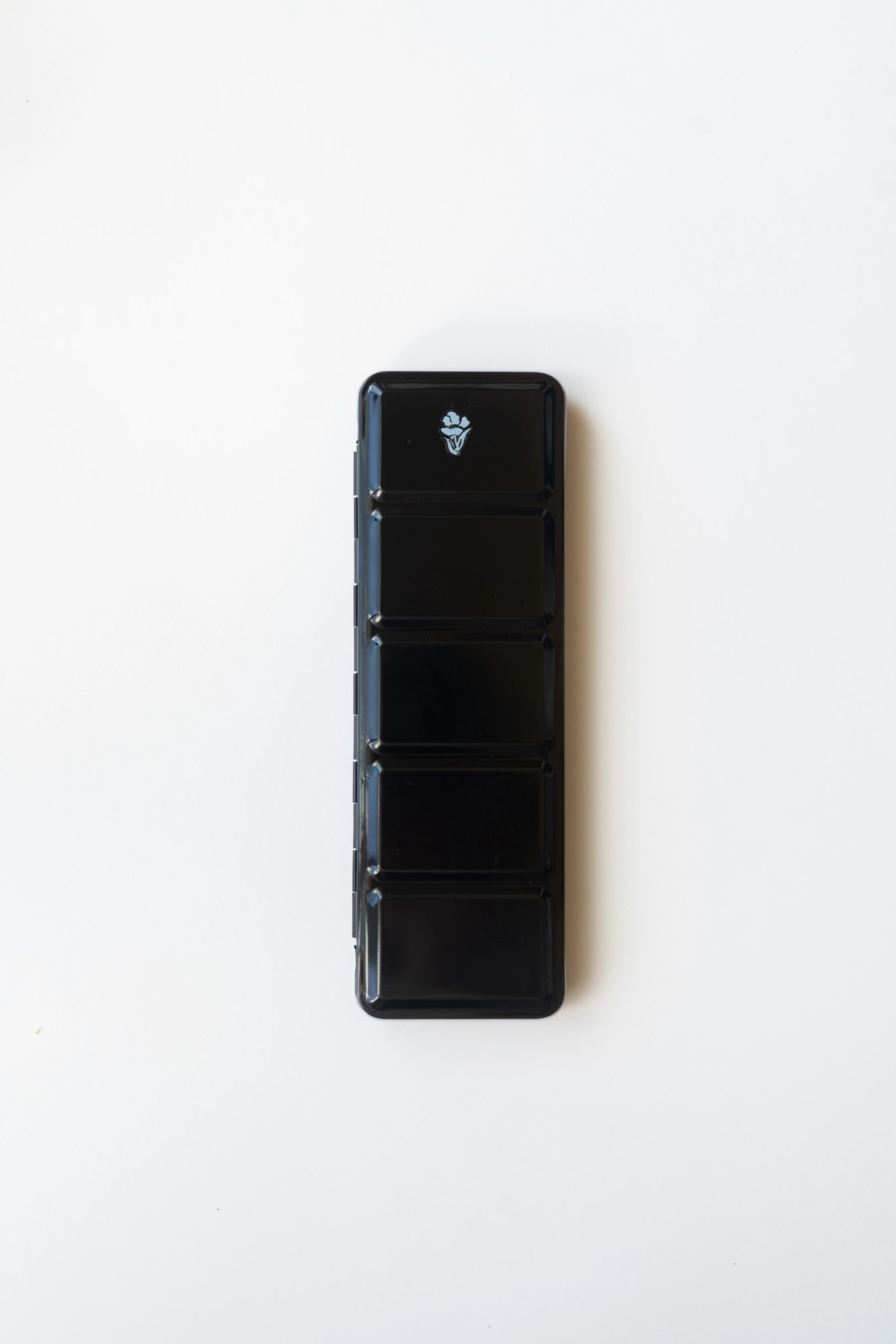 The Faire Watercolor Paint Palette is a black, rectangular palette with five compartments stacked vertically on a white background, resembling a compact pill organizer. The top compartment displays a white water bottle icon.