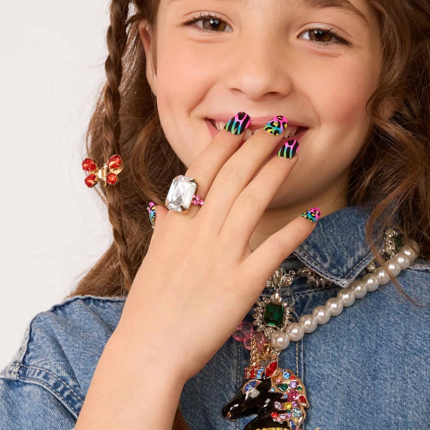 Adorned with the Faire "Days of The Week" press-on nail kit and a stylish clear ring, a young girl in denim beams. Her look features a pearl necklace, a jewel-encrusted horse pendant, and is finished with a playful butterfly hair clip.