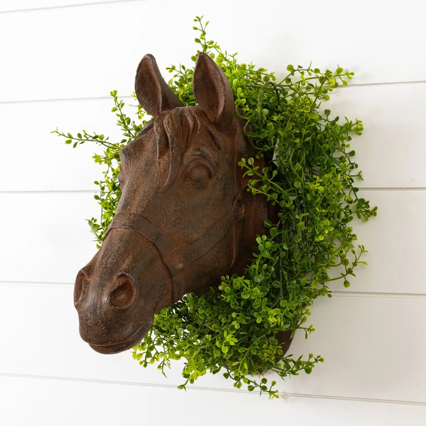 The Faire Horse Head Wall Mount is displayed on a white shiplap wall, accented by a green leafy wreath that forms a decorative frame around the rustic brown sculpture.