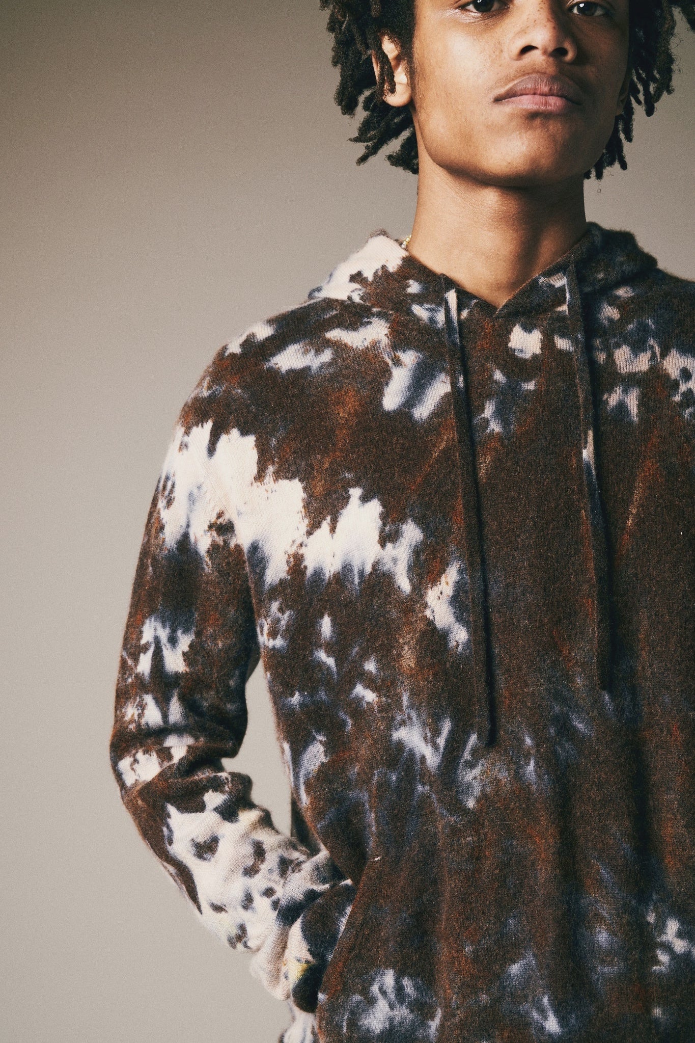 A person wearing the Riverside Tool & Dye Cashmere Hoodie in brown and white, featuring hand-painted details, stands with one hand in their pocket against a neutral background.