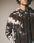 A person wearing the Riverside Tool & Dye Cashmere Hoodie in brown and white, featuring hand-painted details, stands with one hand in their pocket against a neutral background.