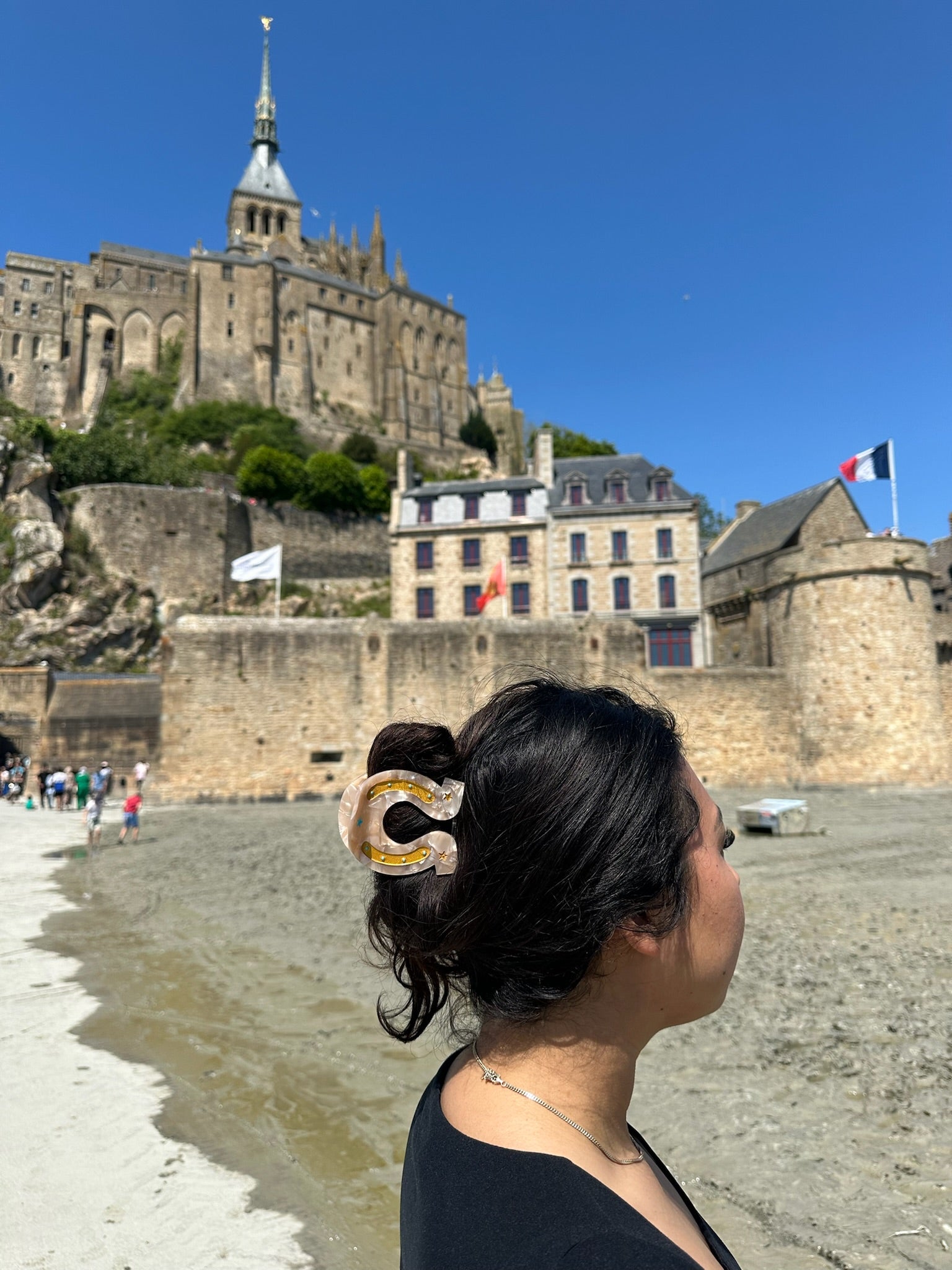 A woman with dark hair in a Faire Fashion Hair Clip stands on sandy ground, looking at Mont Saint-Michel’s historic stone building with flags, under a blue sky.