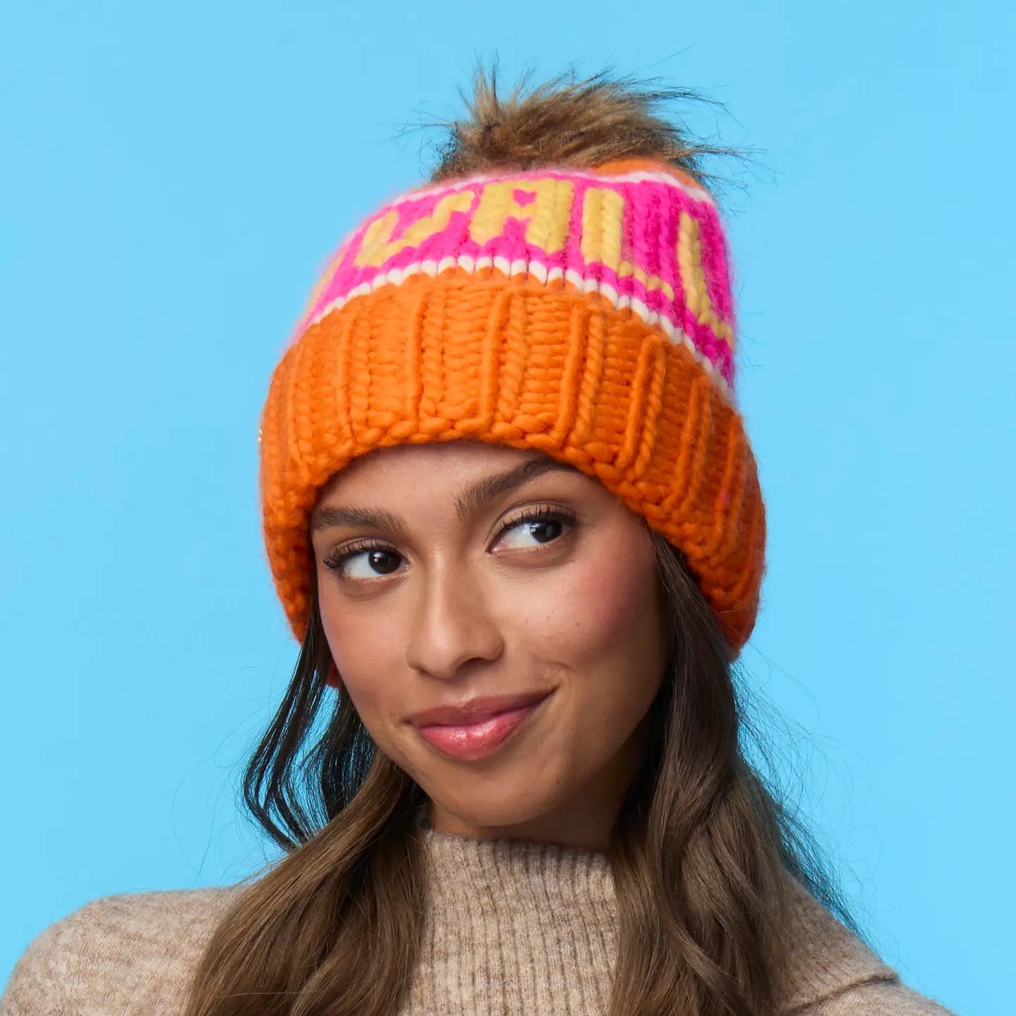 A woman with long brown hair wears a tan sweater and a Faire Knit Ski Hat—an orange luxury knit beanie with "CHILL" in pink and white—against a solid light blue background, smiling softly and looking slightly to the side.