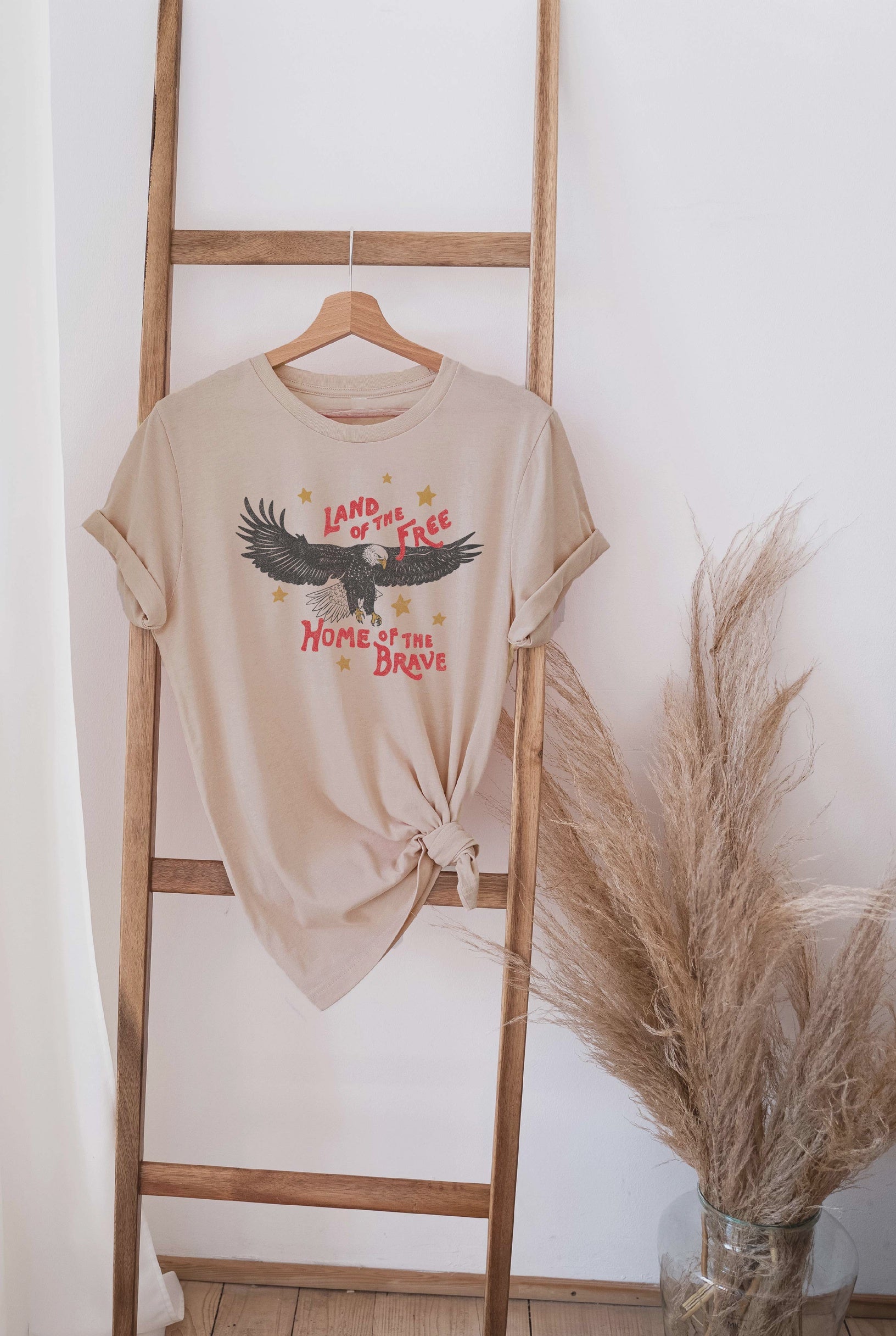 The Oat Collective Land of the Free Home of the Brave T-Shirt, a unisex 100% cotton graphic tee with an eagle and patriotic slogan, hangs on a wooden ladder next to a vase of dried pampas grass.