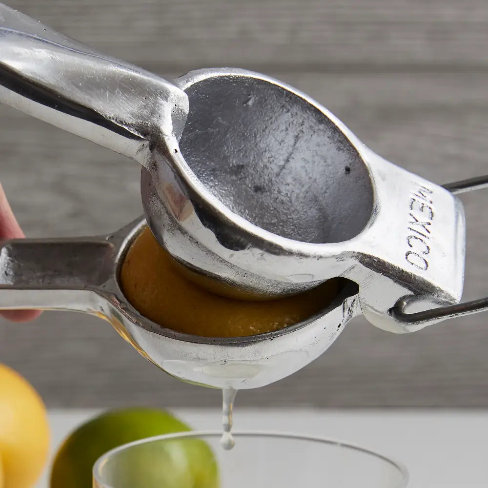 Close-up of the Faire Aluminum Hand Juicer squeezing a lemon, juice dripping into a glass beneath. Blurred in the background are a lime and part of a yellow citrus fruit, illustrating ideal lemon and lime extraction.