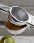 Close-up of the Faire Aluminum Hand Juicer squeezing a lemon, juice dripping into a glass beneath. Blurred in the background are a lime and part of a yellow citrus fruit, illustrating ideal lemon and lime extraction.