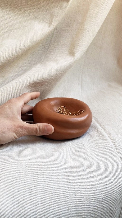 A hand reaches for gold jewelry in Faire’s Sumo Pocket Concrete Bowl—its smooth, round silhouette on a cream fabric backdrop makes it perfect for wabi sabi decor lovers.