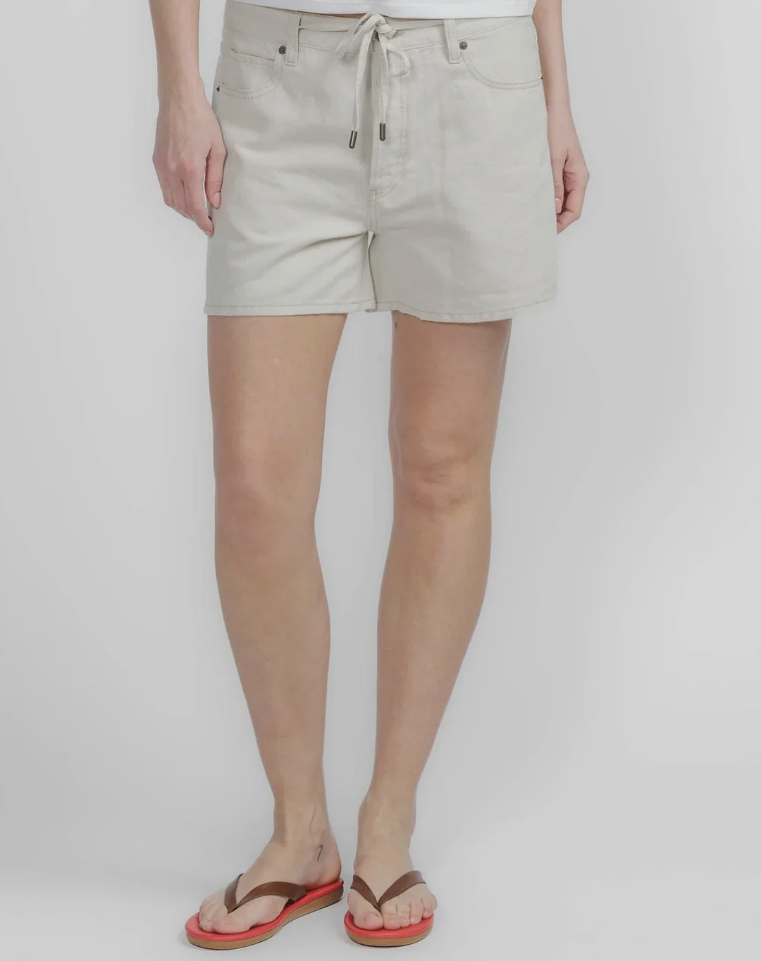 A person wearing FRAME's The Off Duty Long Hang Short, paired with brown flip-flops featuring red soles, stands against a plain light background. The image is cropped at the waist and ankles for a relaxed, effortless vibe.