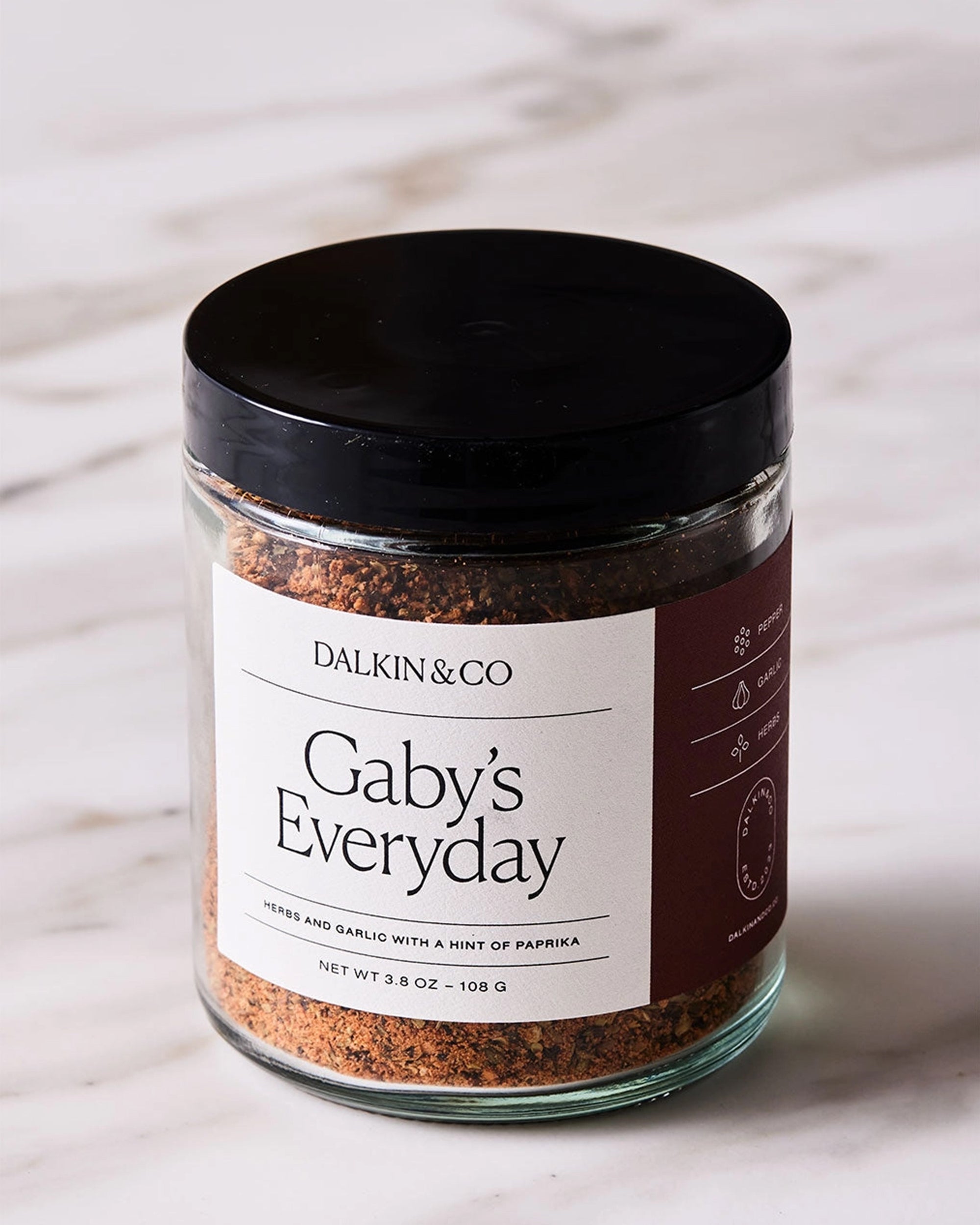 A glass jar with a black lid, labeled &quot;Faire Gaby&#39;s Everyday,&quot; sits on a white marble surface, filled with a flavorful blend of herbs and spices like garlic and smoky paprika.