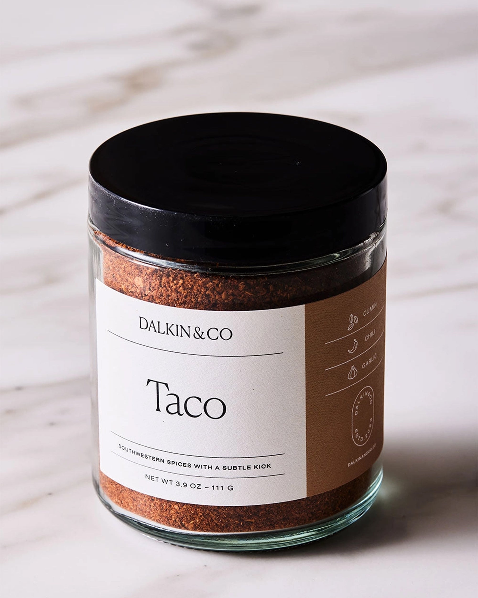 A glass jar with a black lid labeled "Faire Taco Seasoning," filled with Southwestern spices, sits on white marble. The label highlights a subtle kick; net weight is 3.9 oz (111 g).