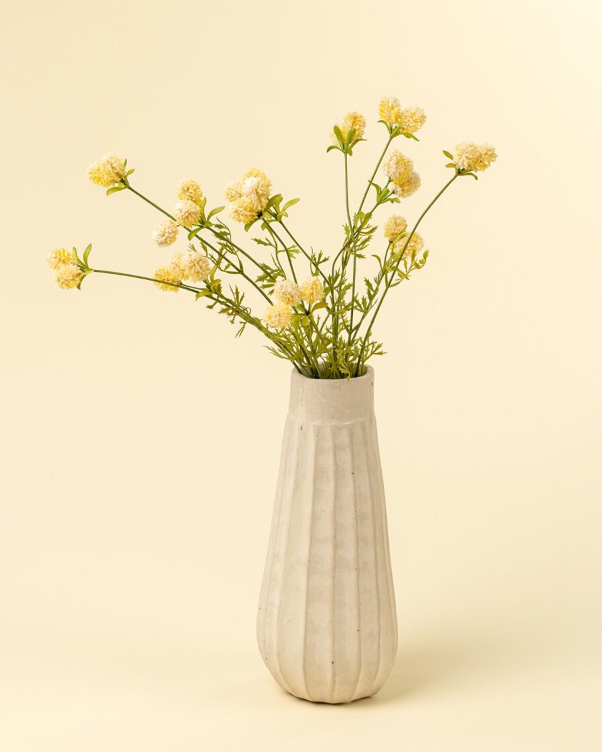The Indaba Dandelion Pom Pom Spray features delicate yellow blooms with thin green stems in a white fluted vase, set against a pale yellow background—an ideal accent decor piece for any room.
