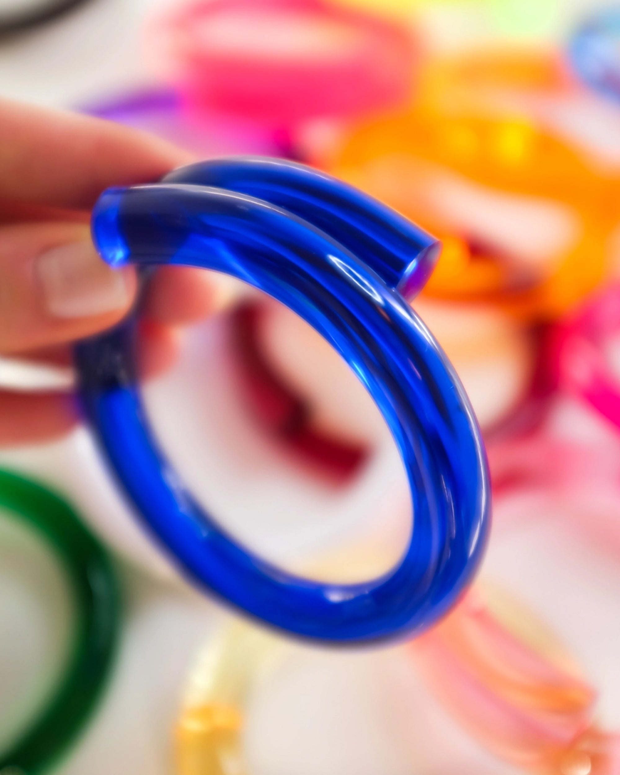 A hand holds the Faire Lucite Dark Blue Neon Bracelet with a twisted design. Blurred pink, orange, green, and yellow bracelets in the background enhance the minimalist jewelry vibe.