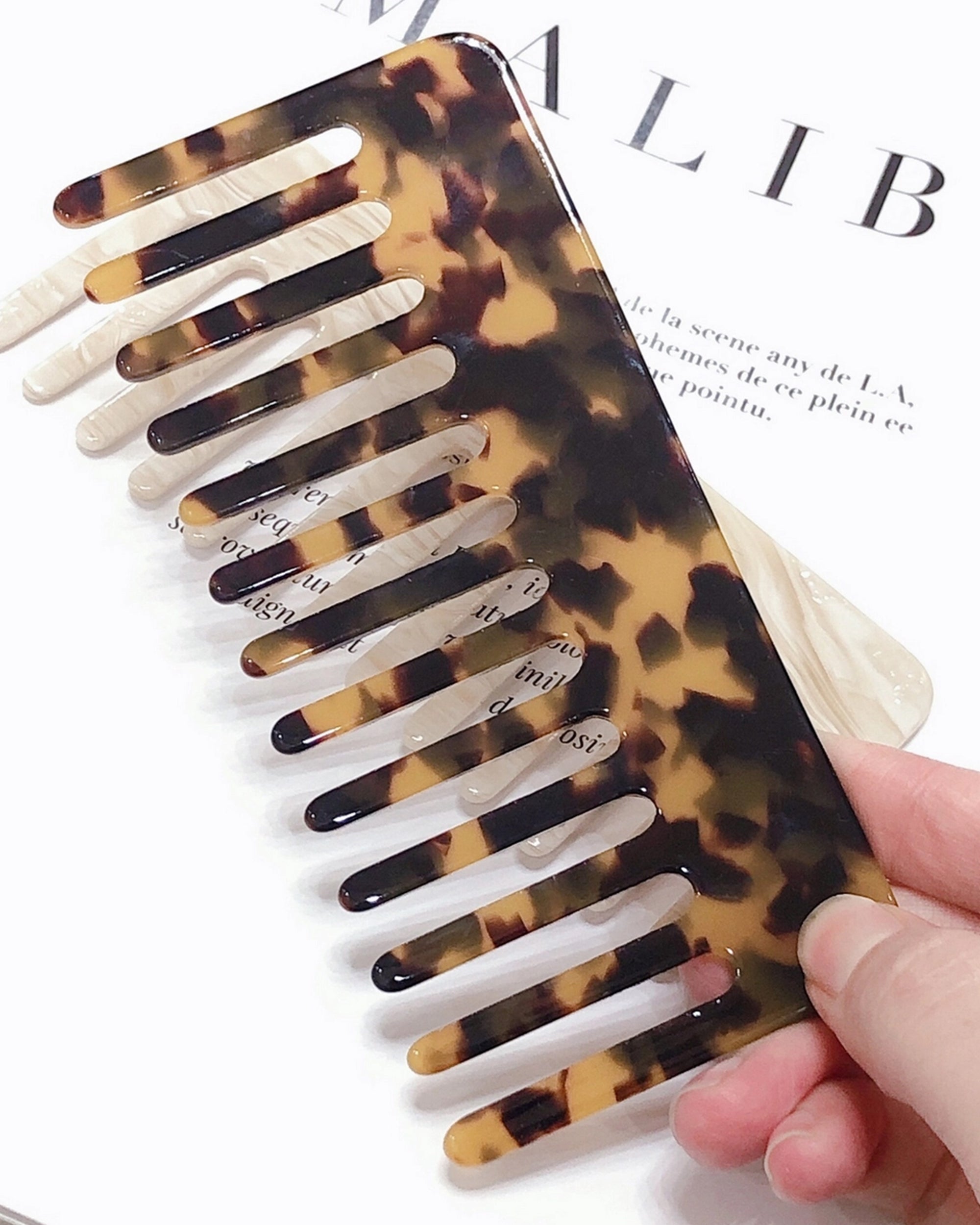 A hand holds the Faire Hair Comb Assorted, featuring a wide-toothed, anti-static design and tortoiseshell pattern, over an open magazine with visible black text in the background.