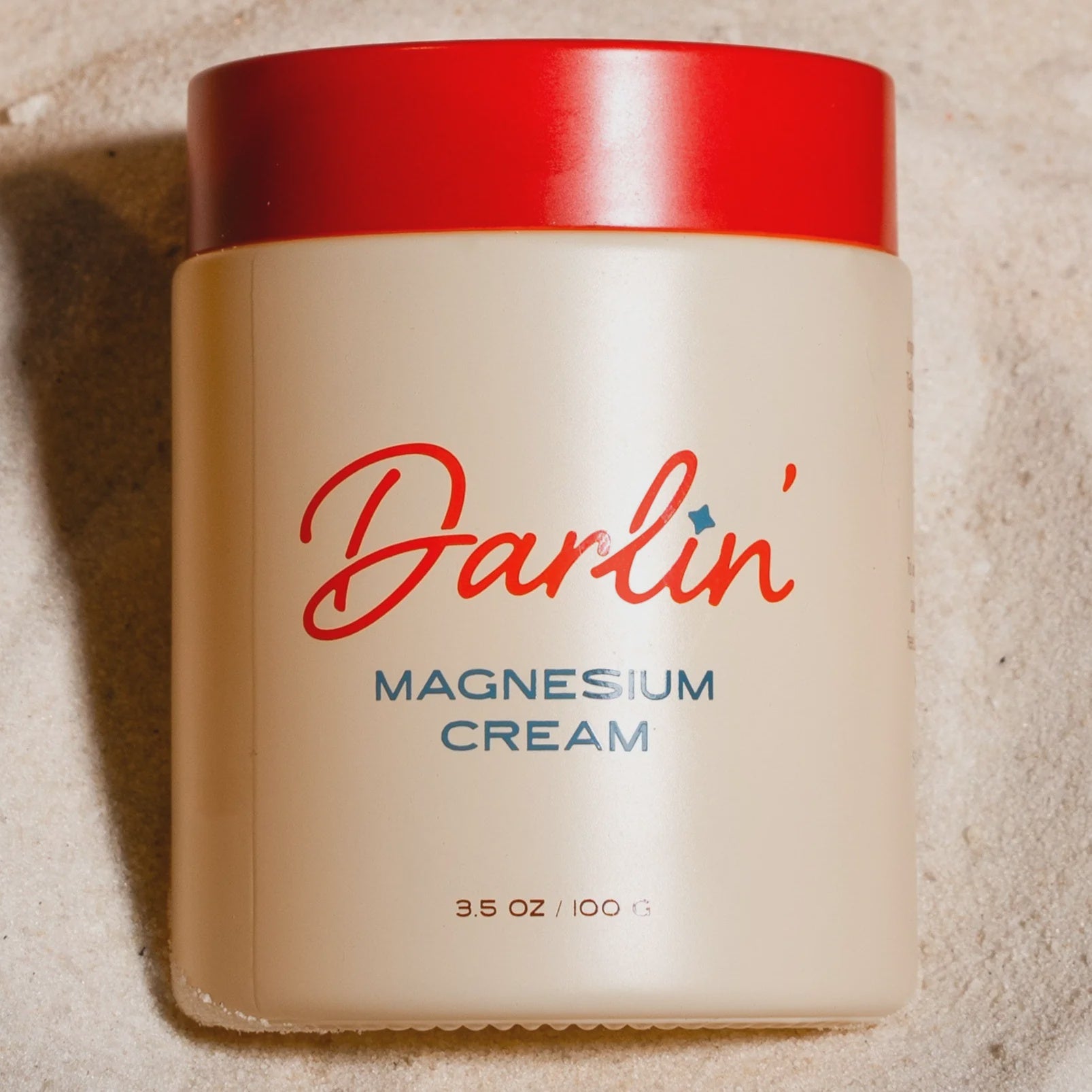 A beige jar of Magnesium Cream with a red lid, labeled "Magnesium Cream 3.5 oz / 100 g," is photographed from above as it rests on sand.