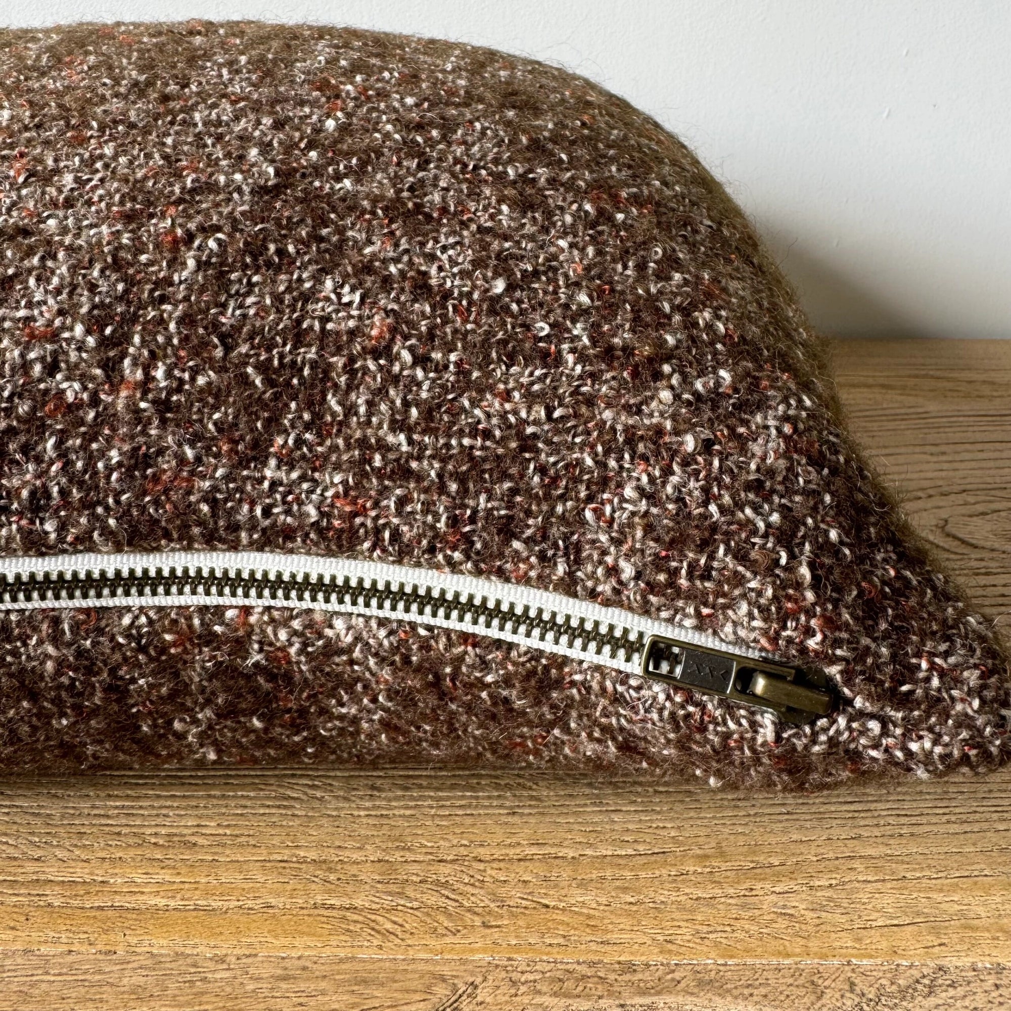 The Faire Adah Coco Wool & Linen Pillow, featuring a textured stonewash finish and visible silver zipper, rests on a wooden surface against a plain light-colored wall.