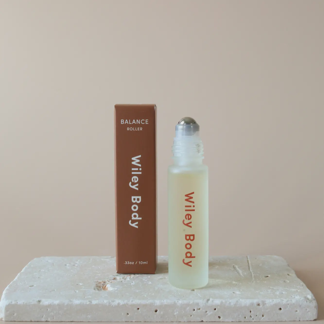 A Wiley Roller, ideal for aromatherapy and wellness, is displayed next to its packaging. The transparent bottle has a metal applicator, and the brown box with white text is placed on a textured stone surface against a calming beige background.