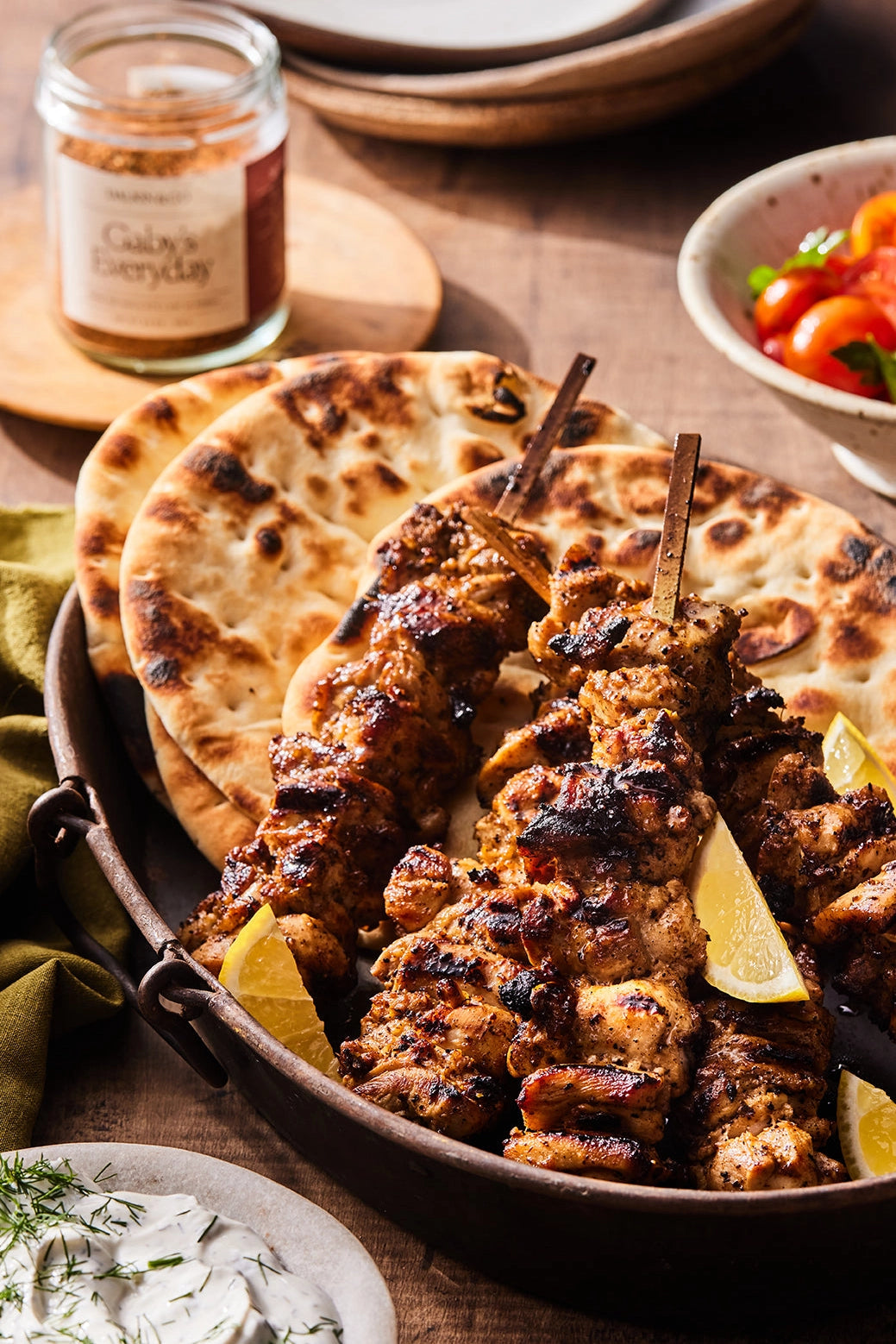 Faire’s Gaby&#39;s Everyday grilled chicken skewers are served with lemon wedges and naan in a rustic bowl, paired with smoky paprika blend, cherry tomatoes, and herbed sauce in the background.