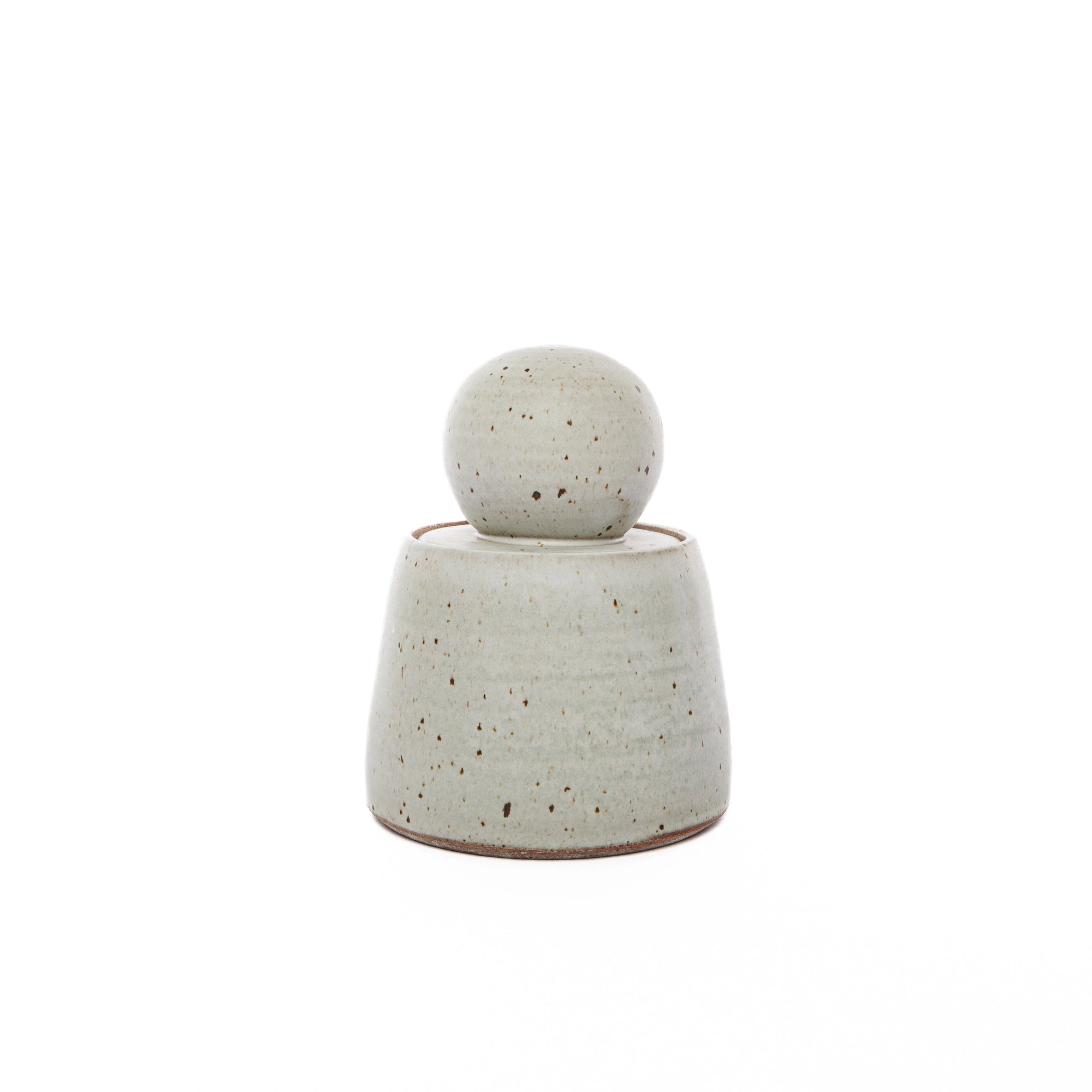 The Faire Alabaster White Stash Jar is a small, handmade stoneware container with a rounded lid. It features a matte light gray glaze with brown speckles, highlighting its minimalist design, and sits on a white background.