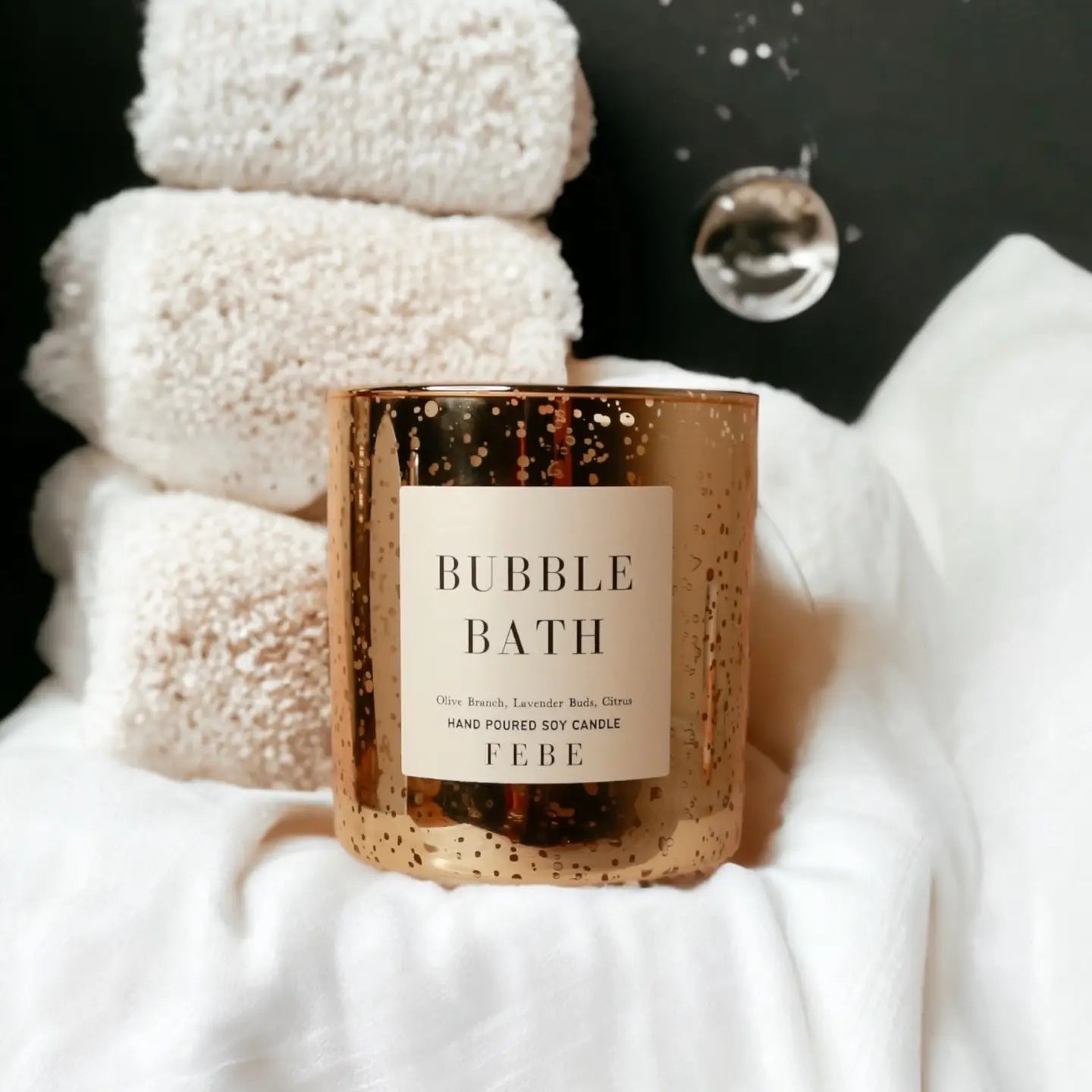 A gold-speckled FEBE Candle from Faire rests on soft, white fabric. Behind it are stacked fluffy towels and a small hanging decoration, evoking a cozy, spa-like atmosphere reminiscent of a luxurious Stage Coach Candle experience.