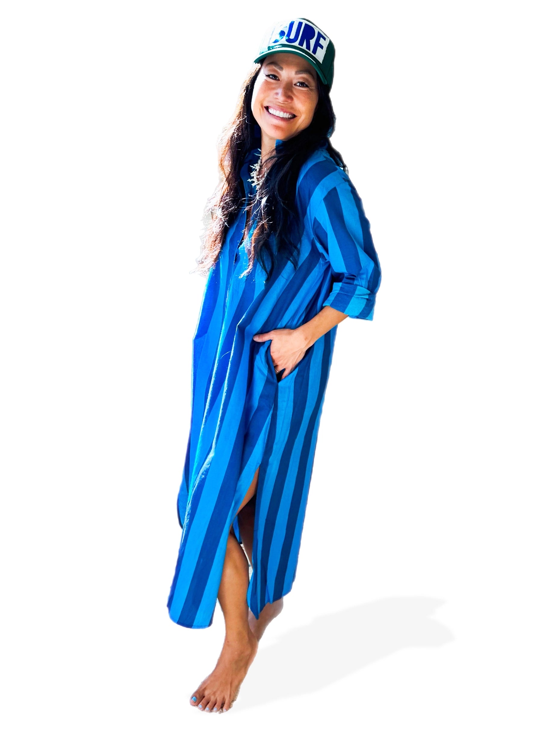 A woman stands barefoot on a white background, wearing the Faire Surf Stripe Shirt Dress and a "SURF" cap, with one hand in her pocket and a smile on her face.