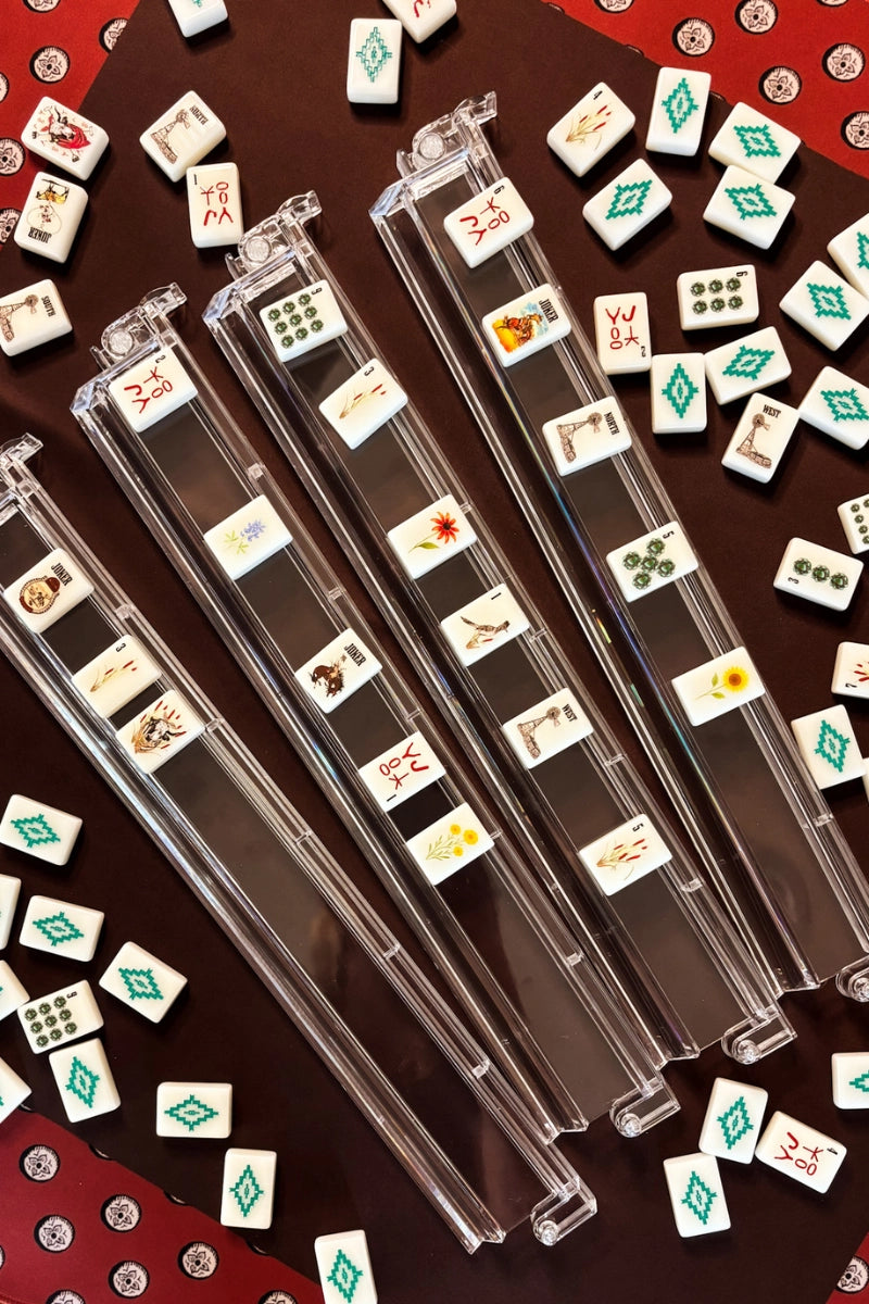 The Faire Wild West Mahjong SET includes four clear racks displaying symbolic tiles, a bag, and mat. Extra tiles are scattered on the dark red mat, hinting at an ongoing or paused game.