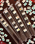 The Faire Wild West Mahjong SET includes four clear racks displaying symbolic tiles, a bag, and mat. Extra tiles are scattered on the dark red mat, hinting at an ongoing or paused game.