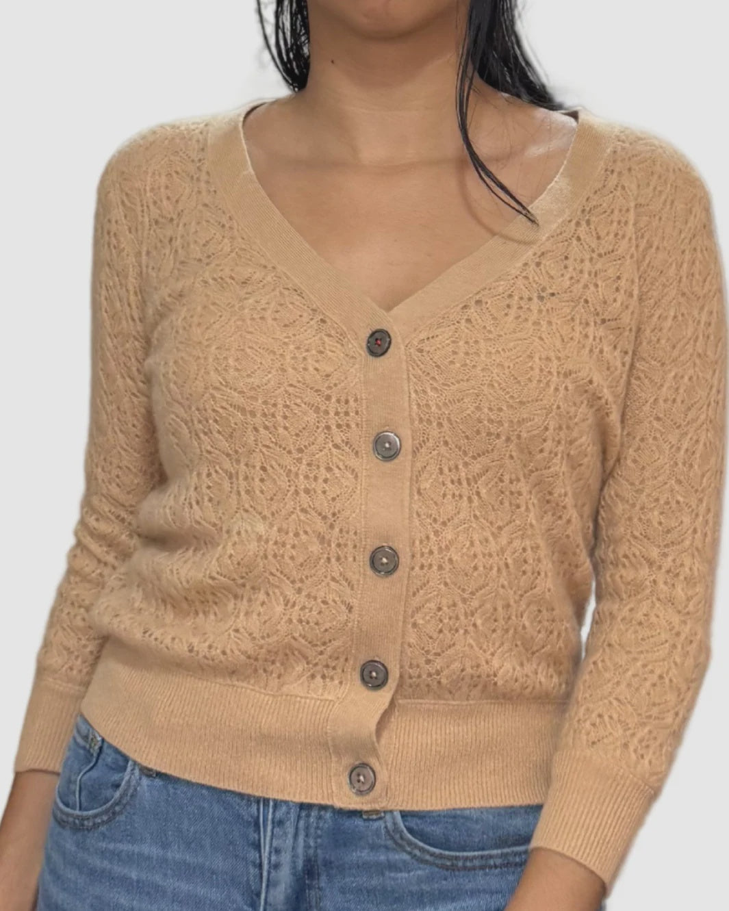 A woman wears the Brazeau Tricot Lace Patch Cardigan—beige with a lacy texture, five front buttons, and three-quarter sleeves—paired with blue jeans, her dark hair draped over one shoulder.