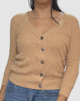 A woman wears the Brazeau Tricot Lace Patch Cardigan—beige with a lacy texture, five front buttons, and three-quarter sleeves—paired with blue jeans, her dark hair draped over one shoulder.