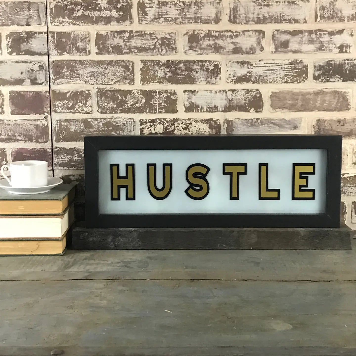 A Faire Lighted Sign with "HUSTLE" in bold black and gold letters rests on a wooden table beside stacked books and a white teacup, all set against a brick wall.