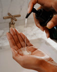 A person dispenses Faire Hand Wash Charlevoix Boreal, a microbiome-friendly liquid soap, from a pump bottle onto their palm over a white sink with a marble countertop and brass faucet in the background.