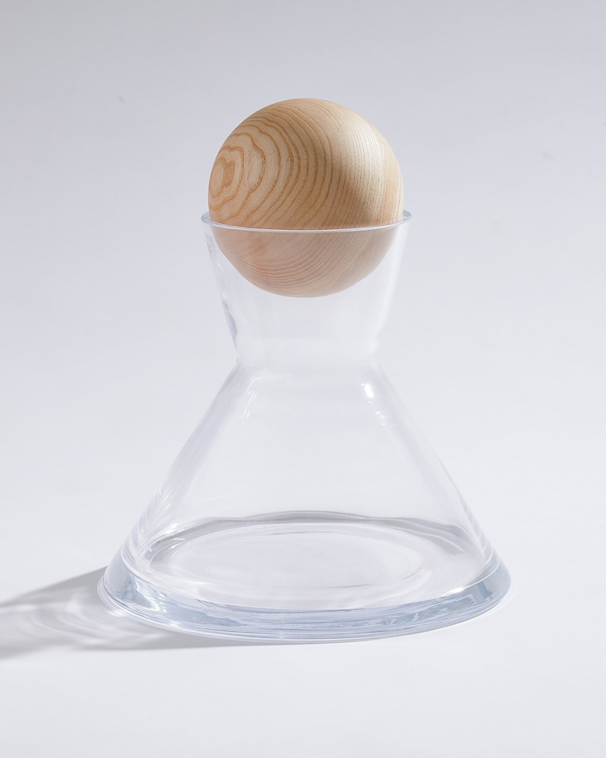 A wooden ashwood sphere stopper tops the clear, hourglass-shaped Global Views 1.11015 Maxwell Decanter L, crafted from hand-blown Portuguese glass and set against a plain white background.