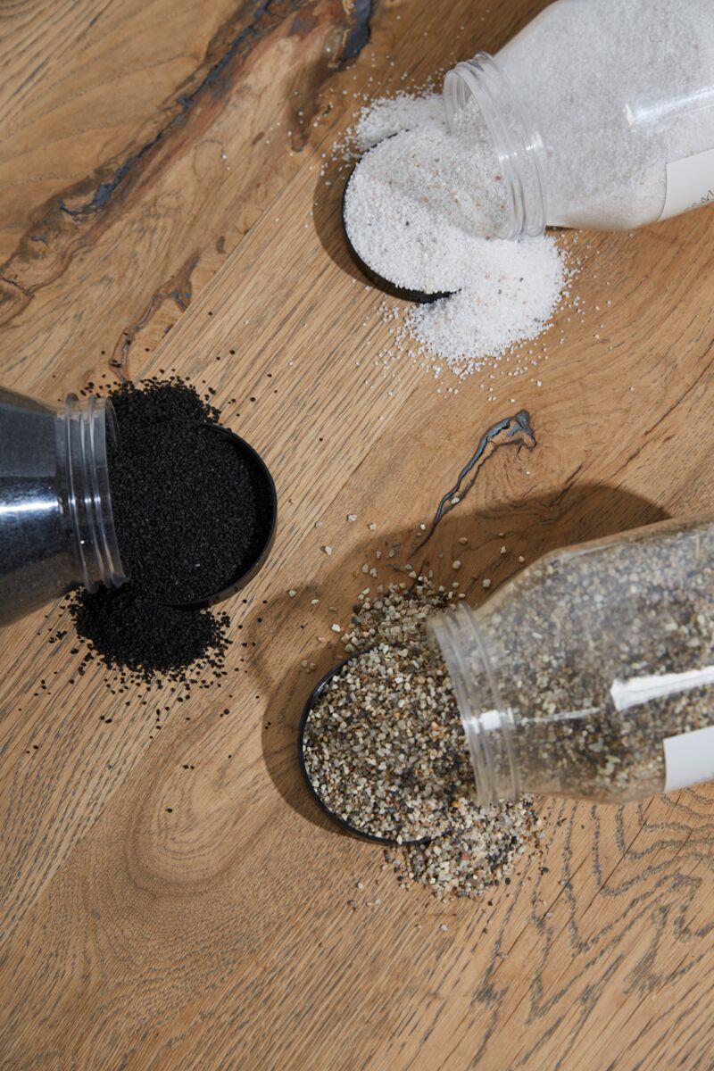 Three Accent Decor Deco Sand containers are tipped over on a wooden surface, each spilling a different type—white salt, black Deco Sand, and a coarse speckled mix—suggesting natural minerals or spices for creative design ideas.