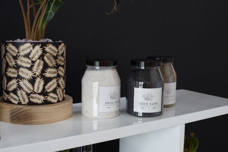 Three Accent Decor "Deco Sand" jars in white, black, and brown sit on a shelf next to a potted plant with a fern leaf pattern, all arranged against a dark wall.