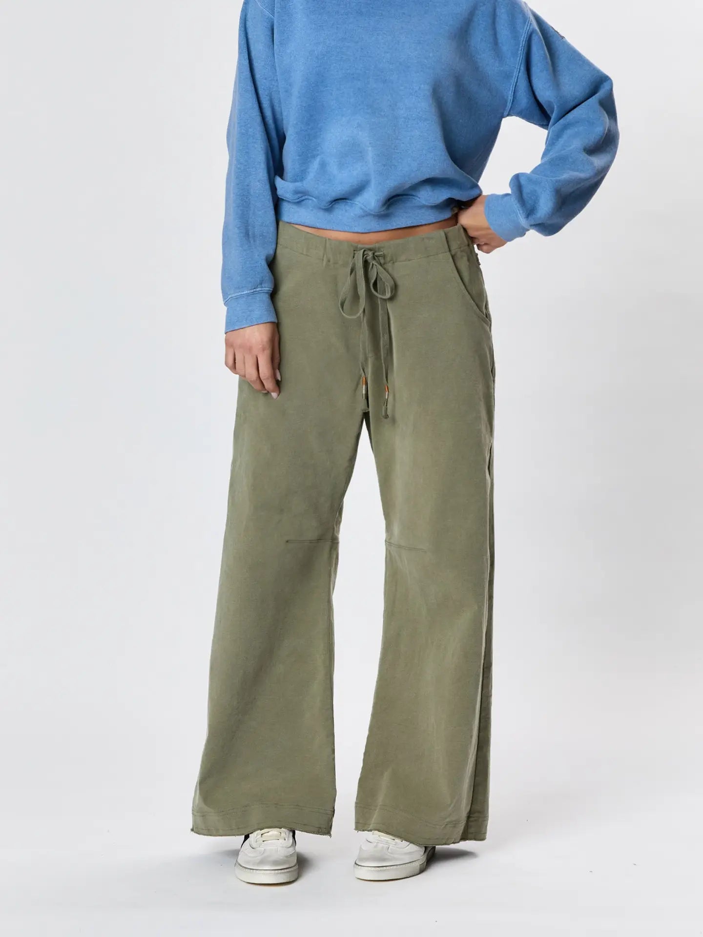 Wearing a blue long-sleeve top, Faire’s Gwen Boyfriend Wide Leg Pant in olive green, and white sneakers, a person stands with one hand on their hip against a plain light background.