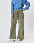 Wearing a blue long-sleeve top, Faire’s Gwen Boyfriend Wide Leg Pant in olive green, and white sneakers, a person stands with one hand on their hip against a plain light background.