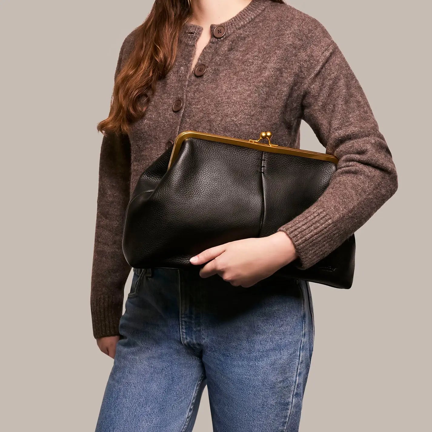 A woman in a brown buttoned sweater and blue jeans holds the Faire Grace Oversized Clutch, a vintage-inspired genuine leather accessory with a gold clasp, against a neutral background.
