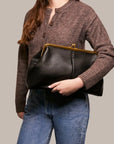A woman in a brown buttoned sweater and blue jeans holds the Faire Grace Oversized Clutch, a vintage-inspired genuine leather accessory with a gold clasp, against a neutral background.