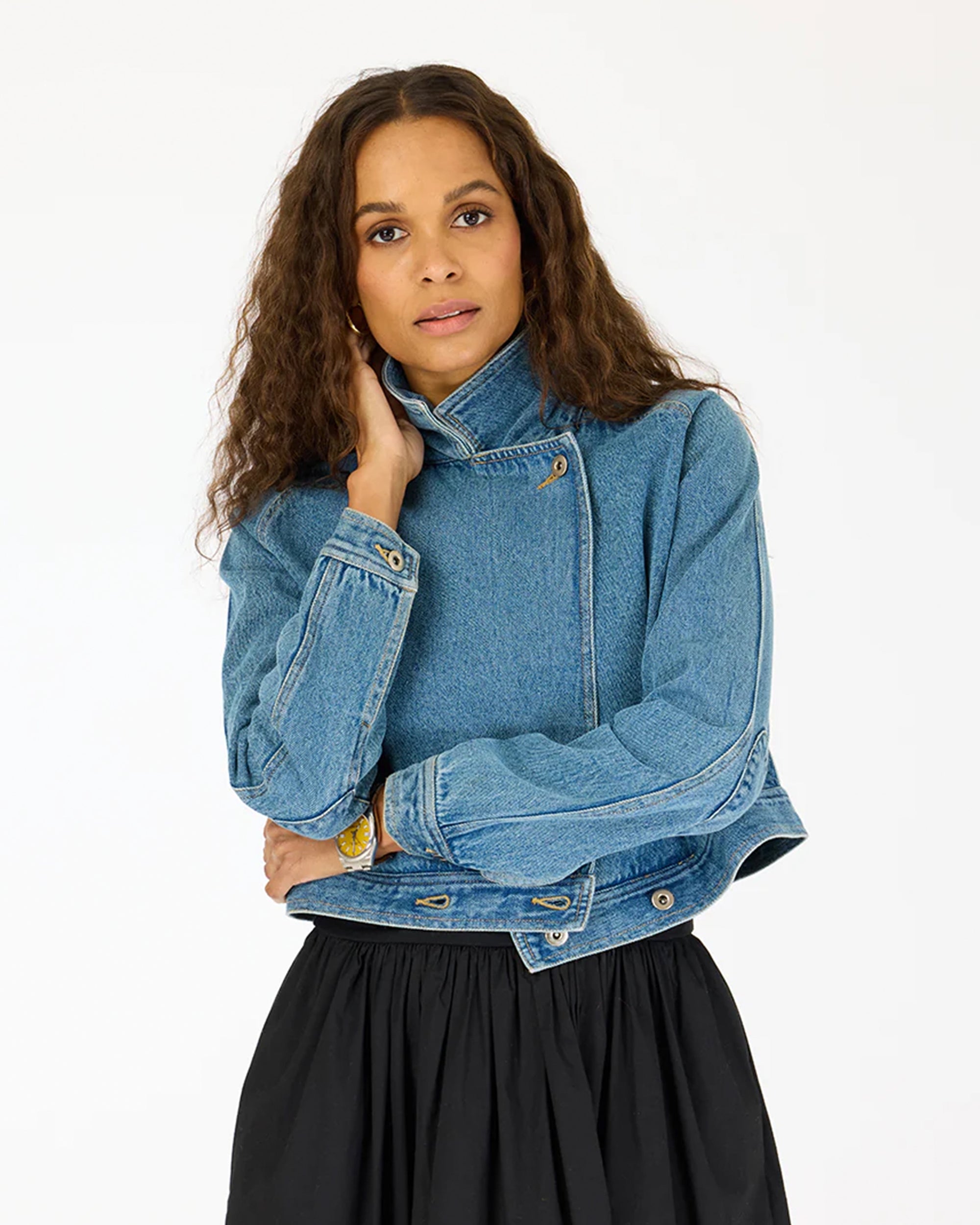 A woman with long curly hair models the Kerri Rosenthal Denim Jacket, featuring heart elbow patches, paired with a black skirt and standing against a plain white background.