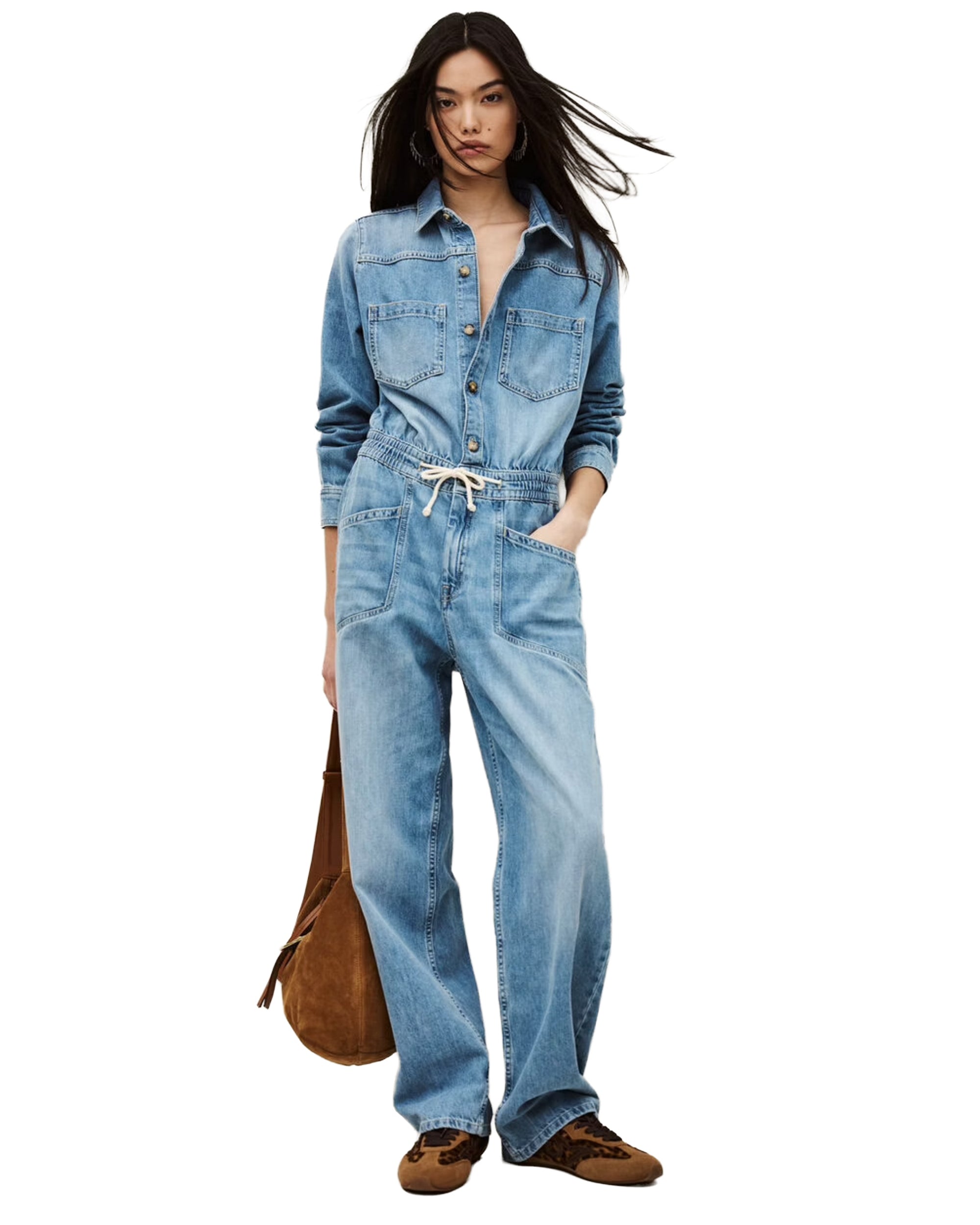 A woman in the BA&SH Jumpsuit Mirella, a light blue denim cargo style with utility pockets and cropped bottoms, paired with brown shoes and a brown suede shoulder bag, stands against a white background.