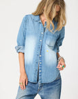 A person with long, wavy blond hair wears the Kerri Rosenthal Denim Shirt Lightweight—an oversized-fit denim top with an embroidered heart—paired with matching jeans. They look down, one hand on their hip and the other at their side.