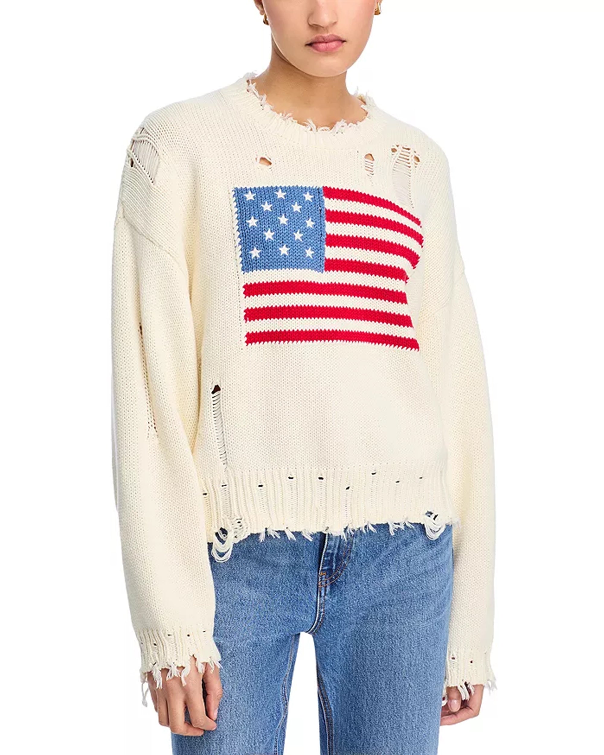 Wearing Denimist’s OVERSIZED CROPPED AMERICAN FLAG SWEATER—an off-white crewneck featuring a large American flag, frayed edges, and distressed holes—paired with blue jeans for a worn-in look.