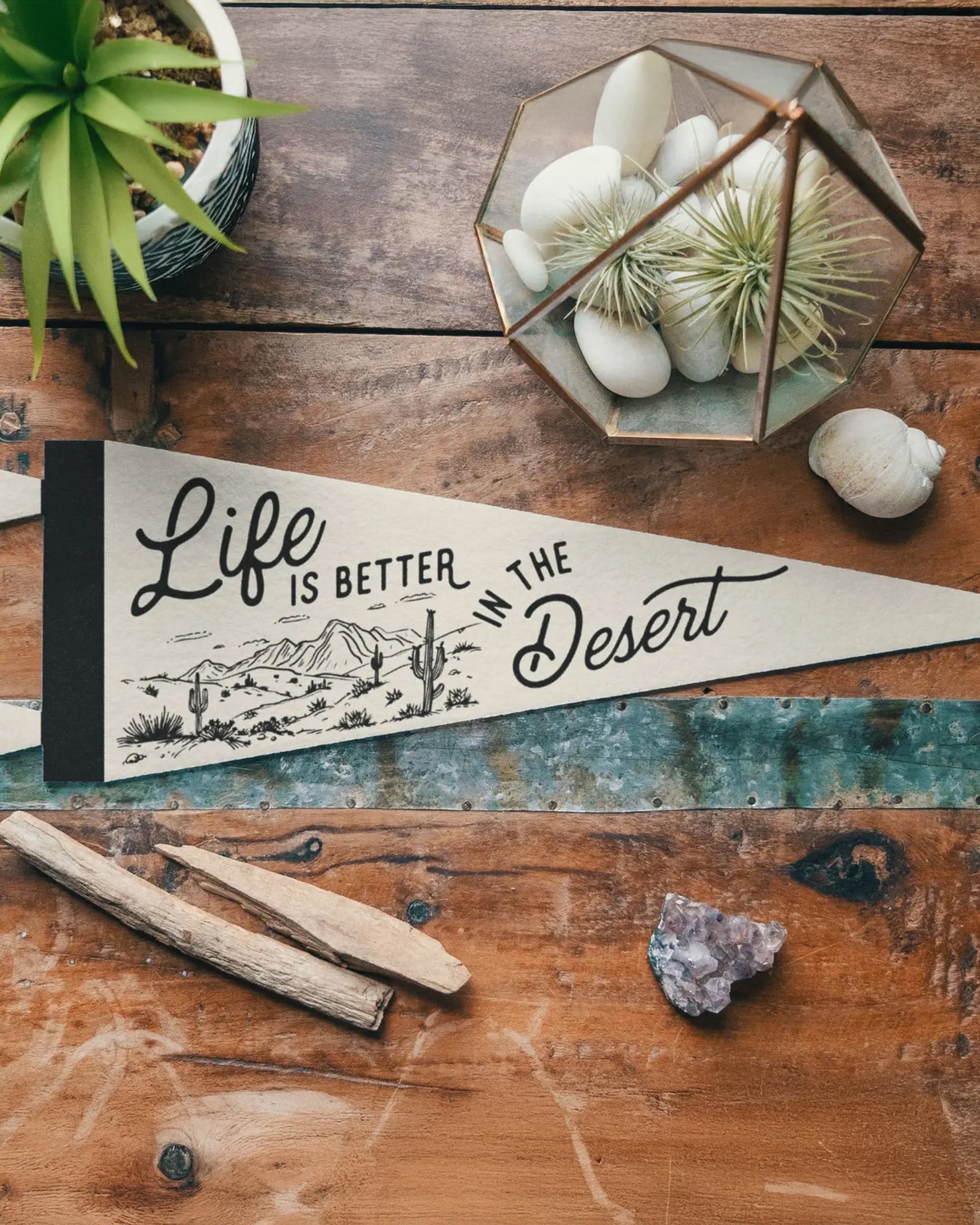 The "Life is Better in The Desert" pennant by Faire features a desert design and sits on a wooden table with a potted succulent, terrarium, wooden sticks, and a purple crystal—ideal for vintage camp banner enthusiasts.