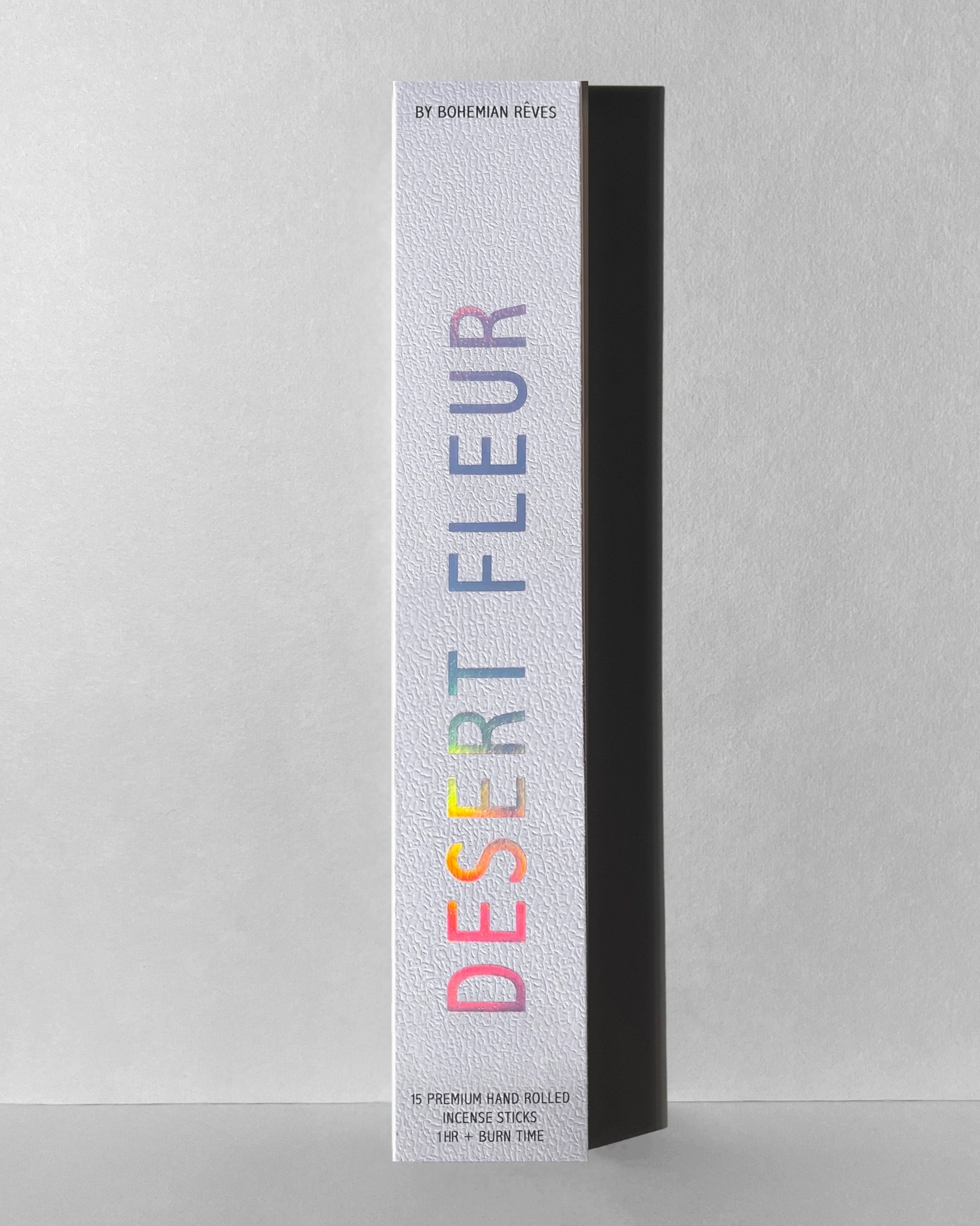 Tall, white rectangular box of Faire Incense Sticks labeled “DESERT FLEUR” in rainbow letters with patchouli notes stands upright on a neutral gray background, casting a shadow to the right.
