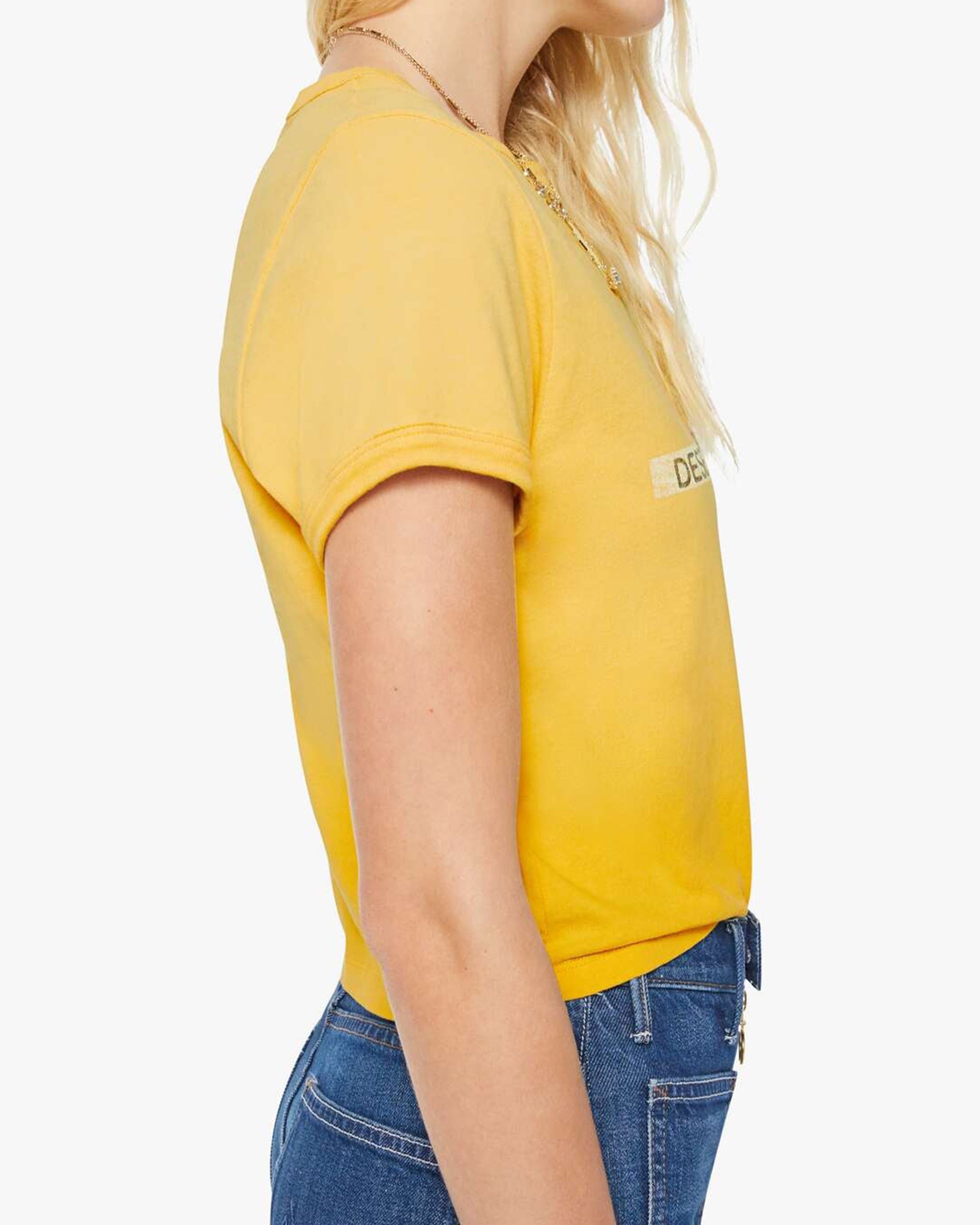 A woman with wavy blonde hair is shown from the side, wearing The Itty Bitty Ringer crop tee by Mother in yellow and blue jeans against a plain white background.
