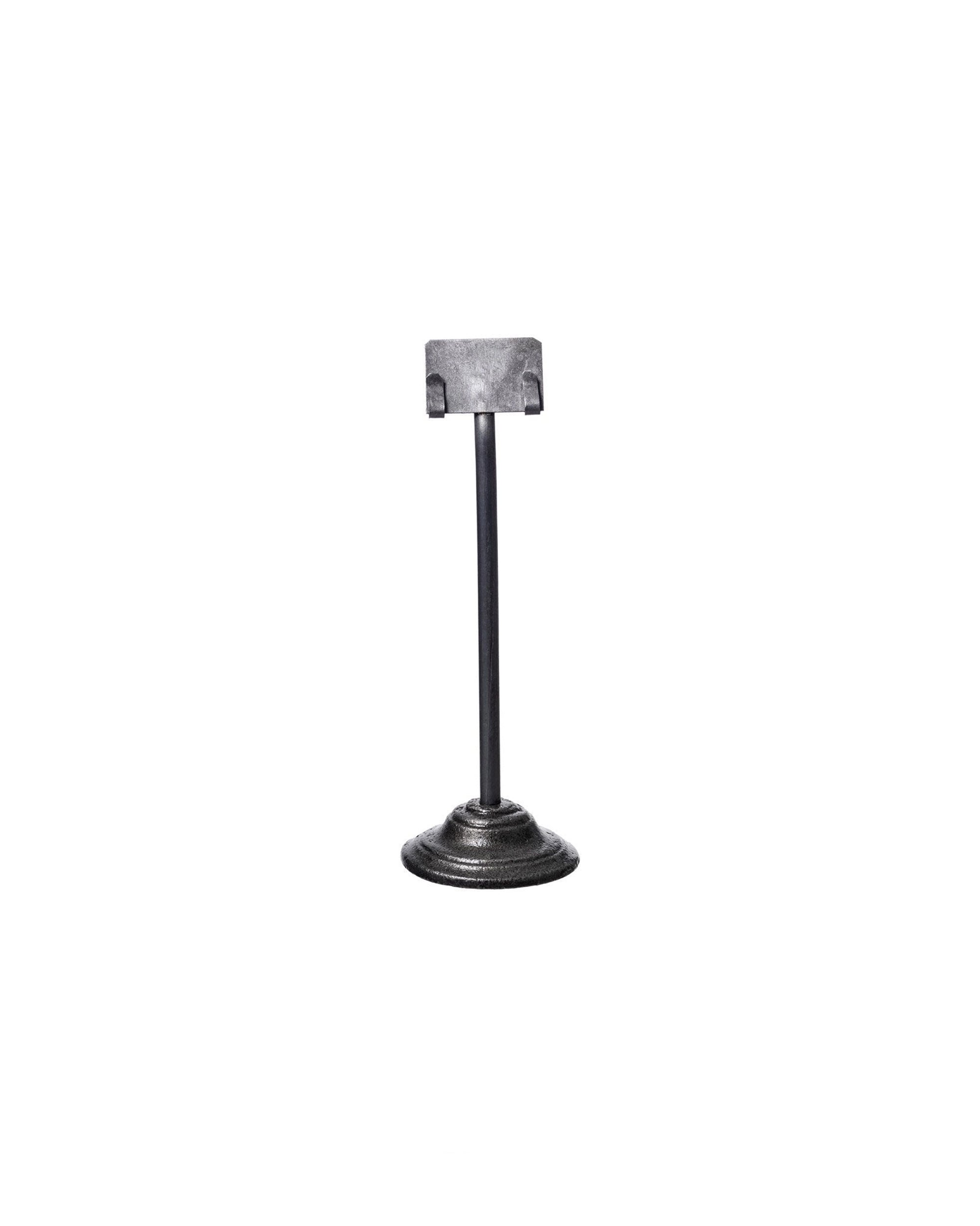 The Faire Desk Card Holder is a small, upcycled steel holder featuring a round, ridged base and a vertical rod with a rectangular clip on top, shown against a plain white background.