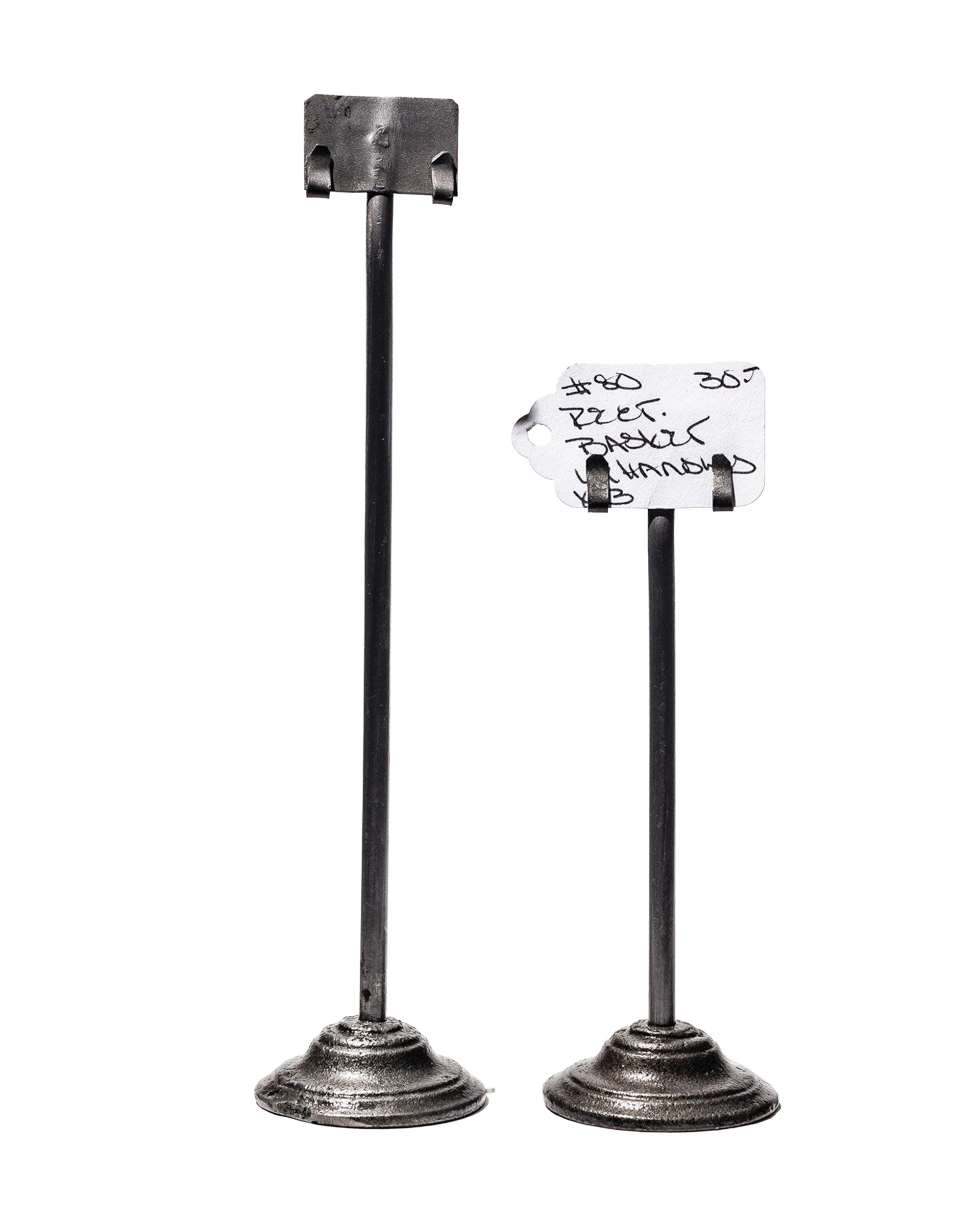 Faire's Desk Card Holder set features two upcycled steel holders made from engine valves. The taller holds a blank metal clip; the shorter secures a handwritten paper tag. Both have round, flat bases.