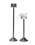 Faire's Desk Card Holder set features two upcycled steel holders made from engine valves. The taller holds a blank metal clip; the shorter secures a handwritten paper tag. Both have round, flat bases.