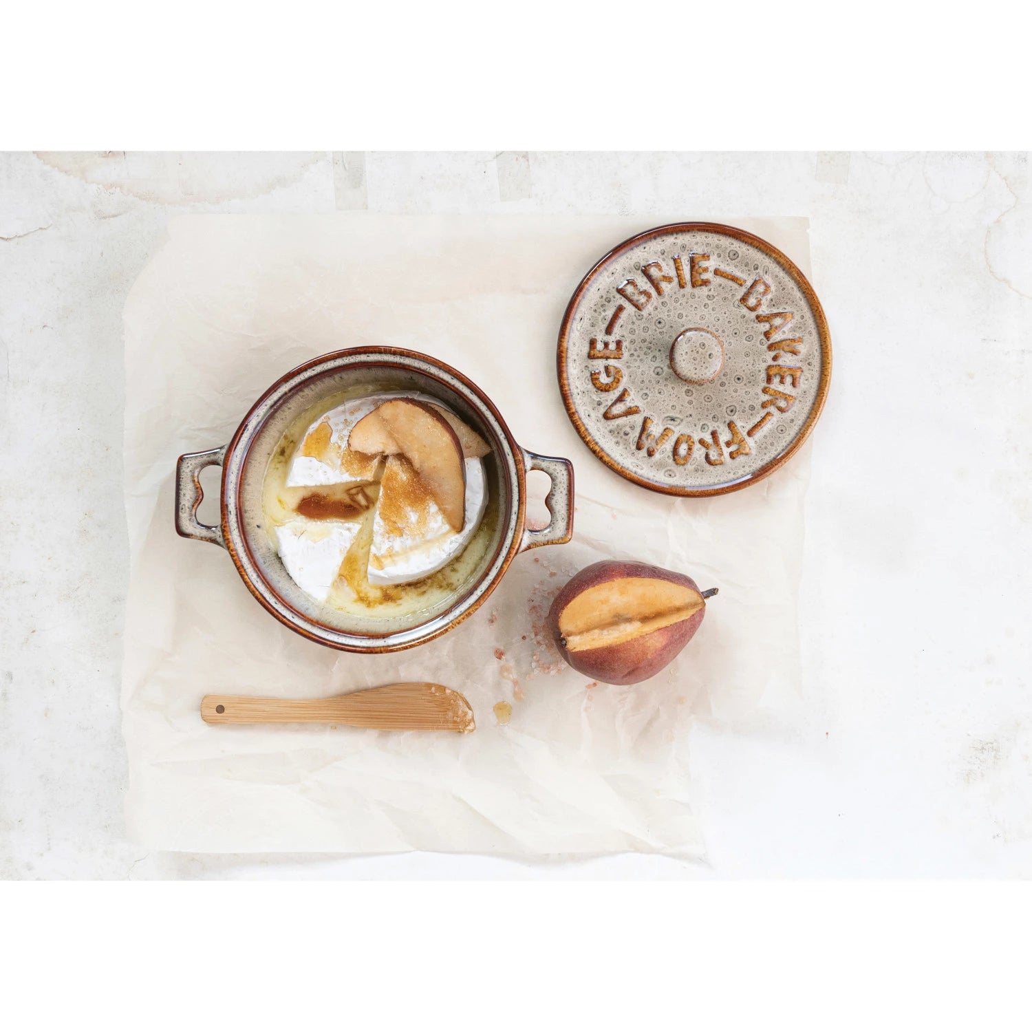 A round Brie cheese melts in a Creative Co-op Brie Baker with "BRIE BAKER" on the lid. Sliced pear tops the cheese, and a halved pear and bamboo spreader rest nearby on white parchment paper.